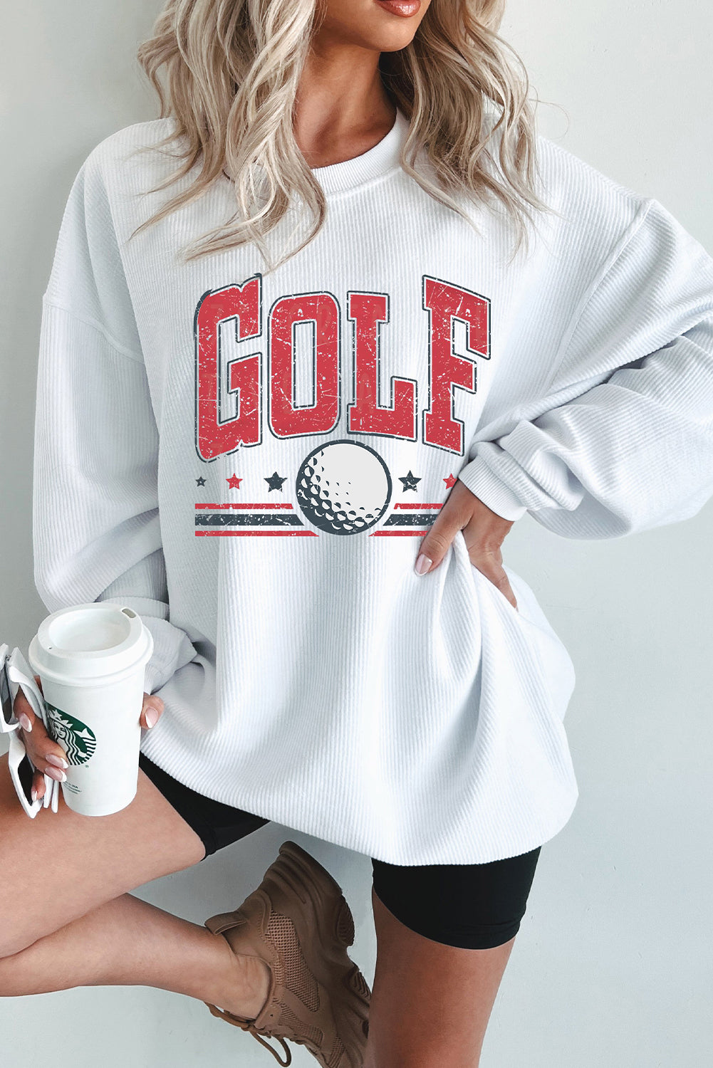 Woman wearing a white sweatshirt with 'GOLF' and a golf ball graphic, holding a Starbucks cup.